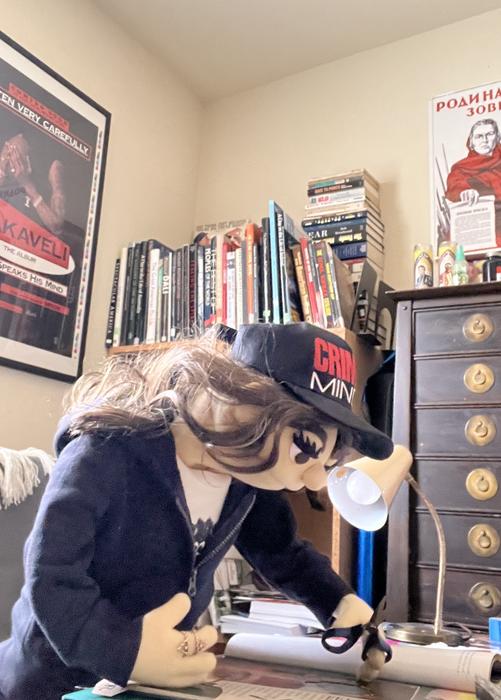 Puppet character with wig and cap at a desk